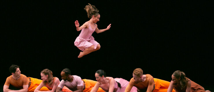 A group of dancers in bright pink and orange costumes lies on their stomachs on a black stage. Above them, Borres, her hair flying, is caught mid-jump, her legs tucked beneath her.