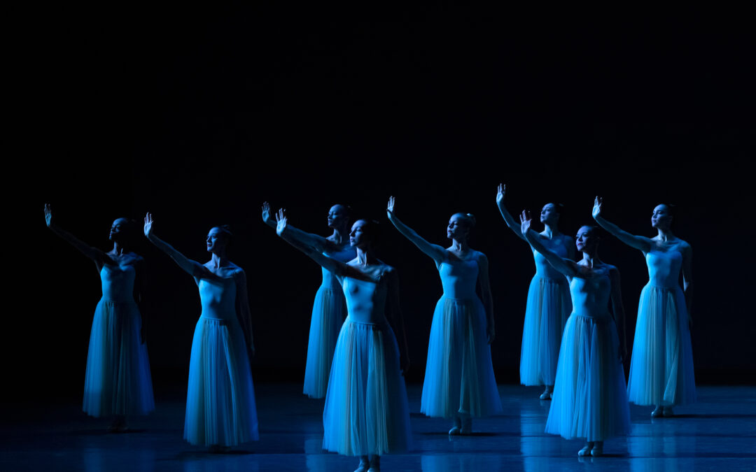 A group of dancers in long, pale blue tutus stands onstage, bathed in blue light, their right arms raised with palms flexed as if to block the sun from their eyes.