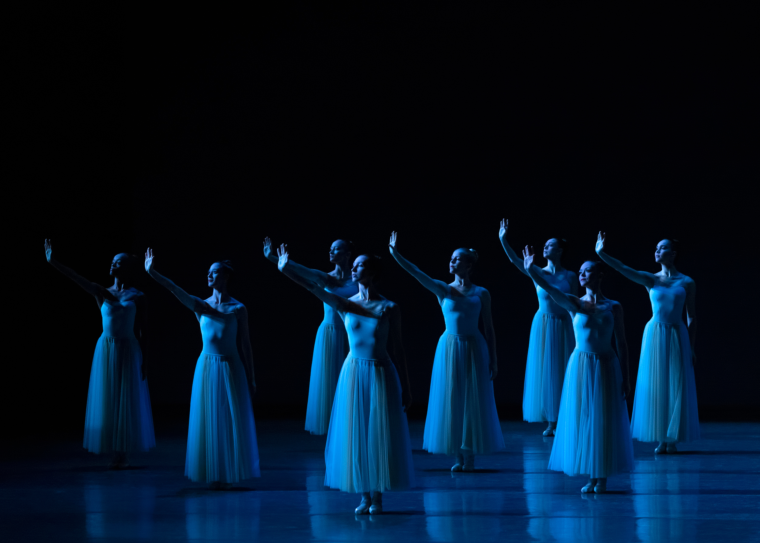 A group of dancers in long, pale blue tutus stands onstage, bathed in blue light, their right arms raised with palms flexed as if to block the sun from their eyes.