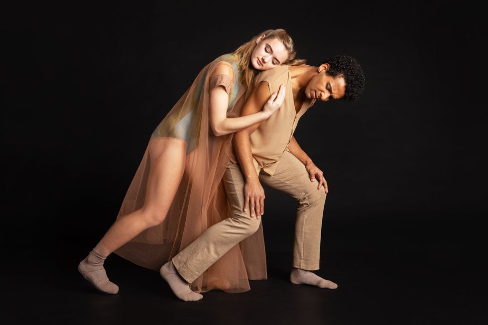 In tan and beige garments, Grace Rookstool rests her forehead on Corey Bourbonniere's shoulder, both of their knees bending as they curve forward.