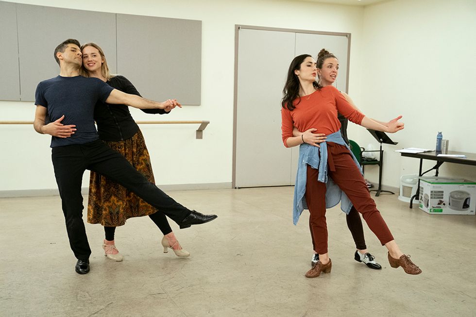 Yazbeck and Pynenburg imitate Sullivan and Dorrance as they show a step in a rehearsal studio. Sullivan, in front, balances on once leg, supported by Dorrance holding her waist and hand from behind.
