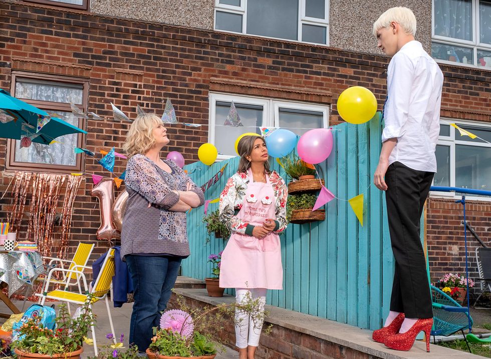 A young man in a rumpled dress shirt and slacks balances in sparkling ruby heels, looking down as he speaks to two older women in casual dress. Around the back garden are balloons and streamers for a birthday party.