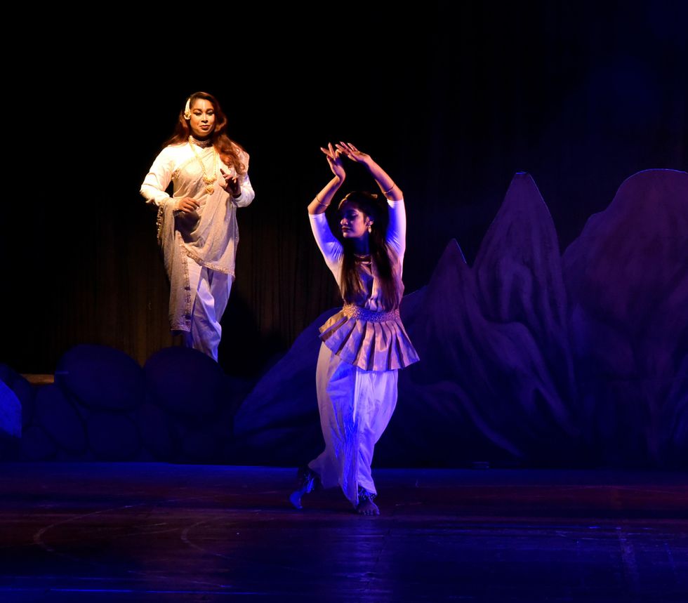 A dancer poses on a shadowy stage, eyes downcast as she brings the backs of her hands together over her head. Upstage, a well lit woman in white observes with an almost-smile, a hand rising as though to gesture to her.