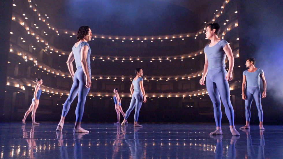 Six dancers in simple grey blue costumes and ballet slippers stand onstage, the empty house visible beyond them, its lights reflecting on the marley.