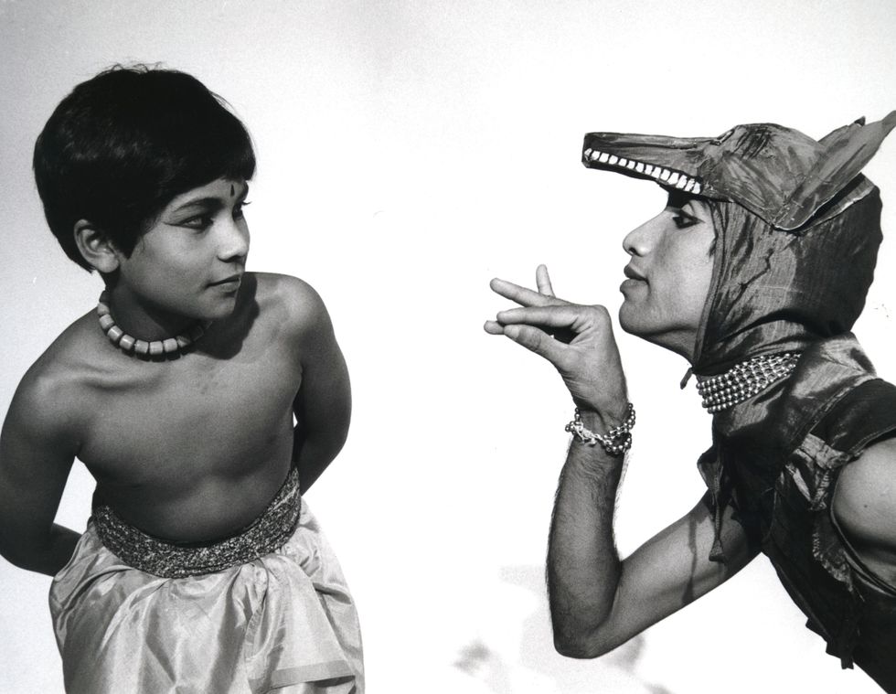 In a black and white archival image, a young Akram Khan poses with his hands behind his back, looking uncertainly at an older dancer costumed as an animal and gesturing to him with a delicate mudra.