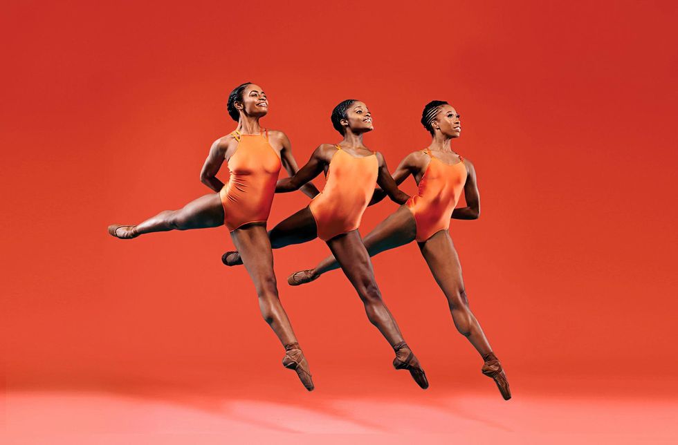 Against an orange-red background, three Black women ballet dancers in orange leotards and flesh tone pointe shoes leap together. Their arms are behind each other's backs as they temps levu00e9 in low arabesque, smiling with their chins lifted to the corner.