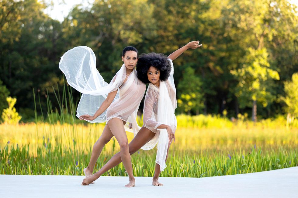 Donna Crump and Kayla Collymore look intently at the camera as they pose on a white stage against a backdrop of sunlit grass and trees. Diaphanous white fabric billows and drapes around them.
