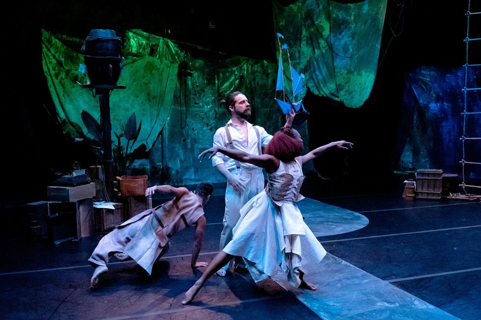 On a shadowy stage set, three dancers in deconstructed dresses with corsets and partial suits with suspenders move separately in a cluster. A white man stands proudly, holding a model ship. A dark skinned woman flows through pliu00e9 in front of him, arms extended lightly. A third dancer goes to the floor, back to the viewer.
