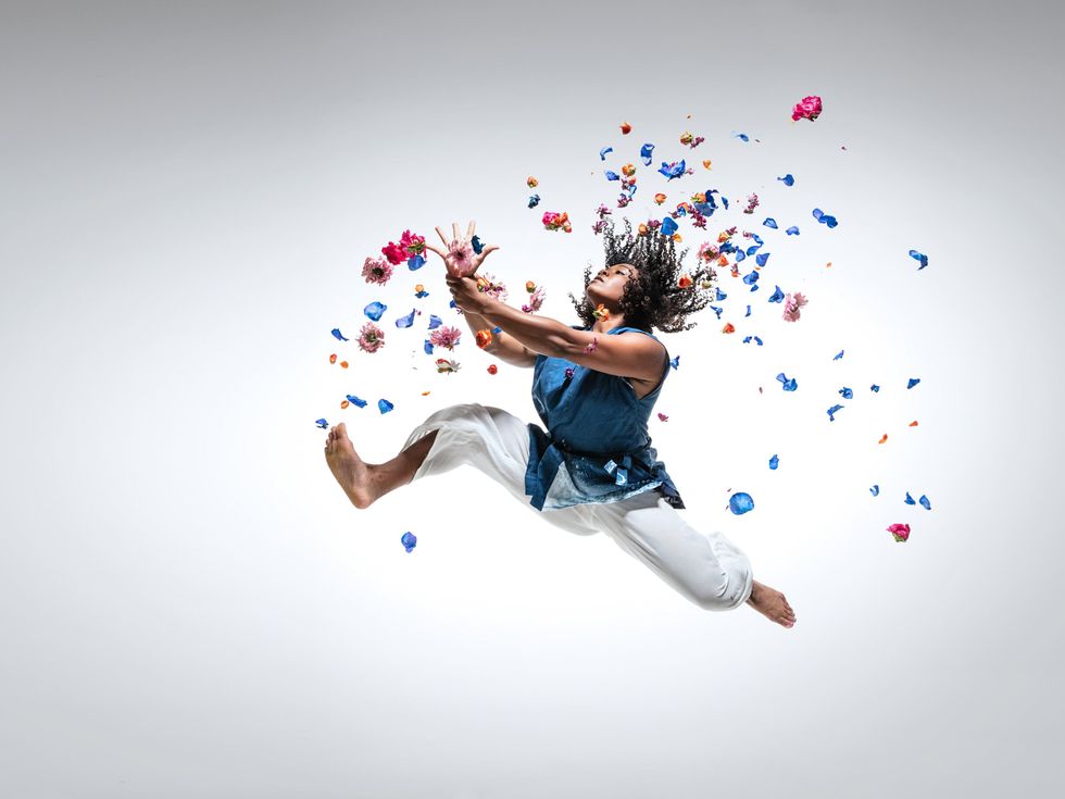 Against a white backdrop, a Black dancer performs a stag leap with flexed feet, holding one wrist before her with the opposite hand, head tipped back. Blue and pink flowers fly and fall around her.