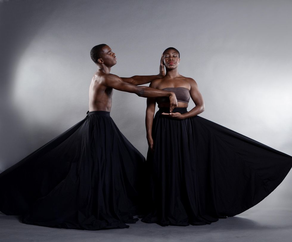Two Black dancers pose against a grey background, both in voluminous black skirts. One faces the camera with eyes serenely closed, one hand cupped at her waist. The other faces her, pressing one hand against her cheek, the other hand extended so his fingertips rest in her palm.