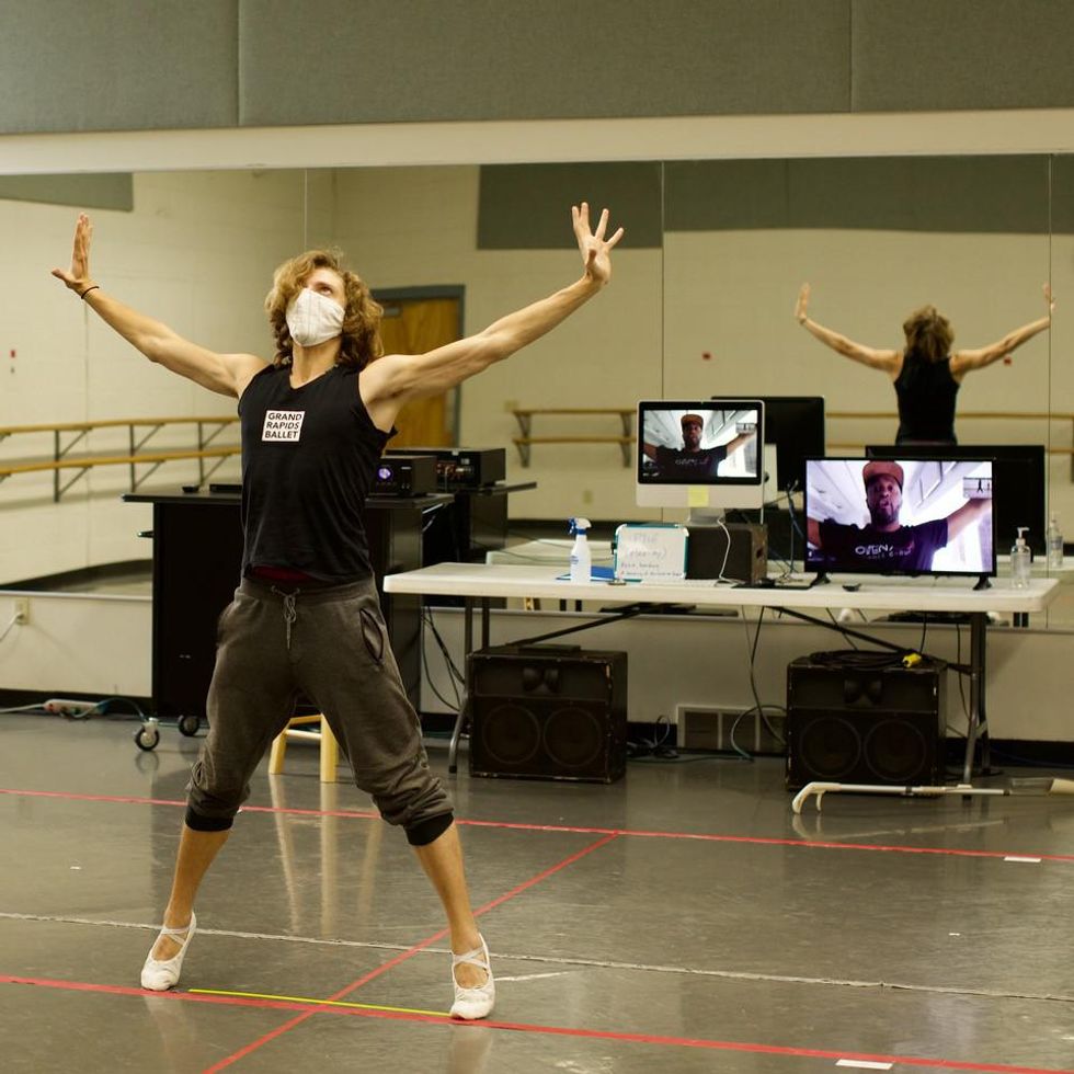 A white male dancer in rehearsal wear poses in parallel second position relevu00e9, arms pressing out. At the front of the room, two screens showing a choreographer demonstrating on video call rest on a table.