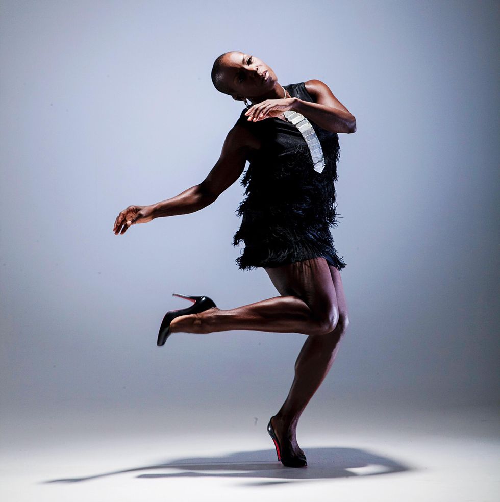 Hope Boykin, a dark-skinned Black woman with a shaved head, hearing wearing high heels and a black cocktail dress, balances on one leg, head gently tipped toward her popped foot.