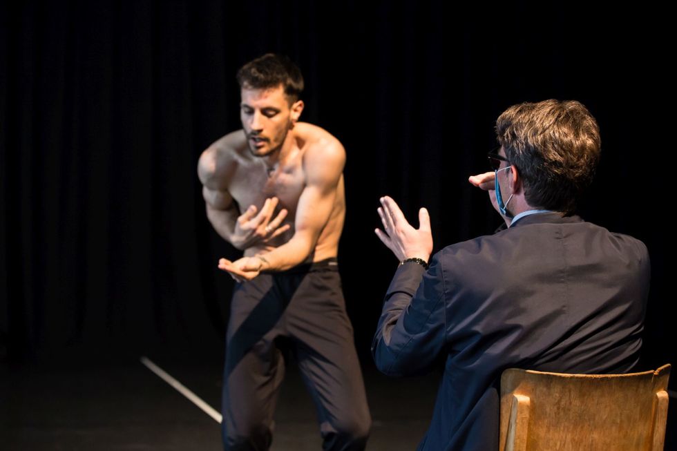 Goecke is seen from behind, seated as he gestures with his hands while he watches the dancer in front of him. The shirtless male dancer contracts, fingertips to his sternum as the other hand extends palm up at his waist.