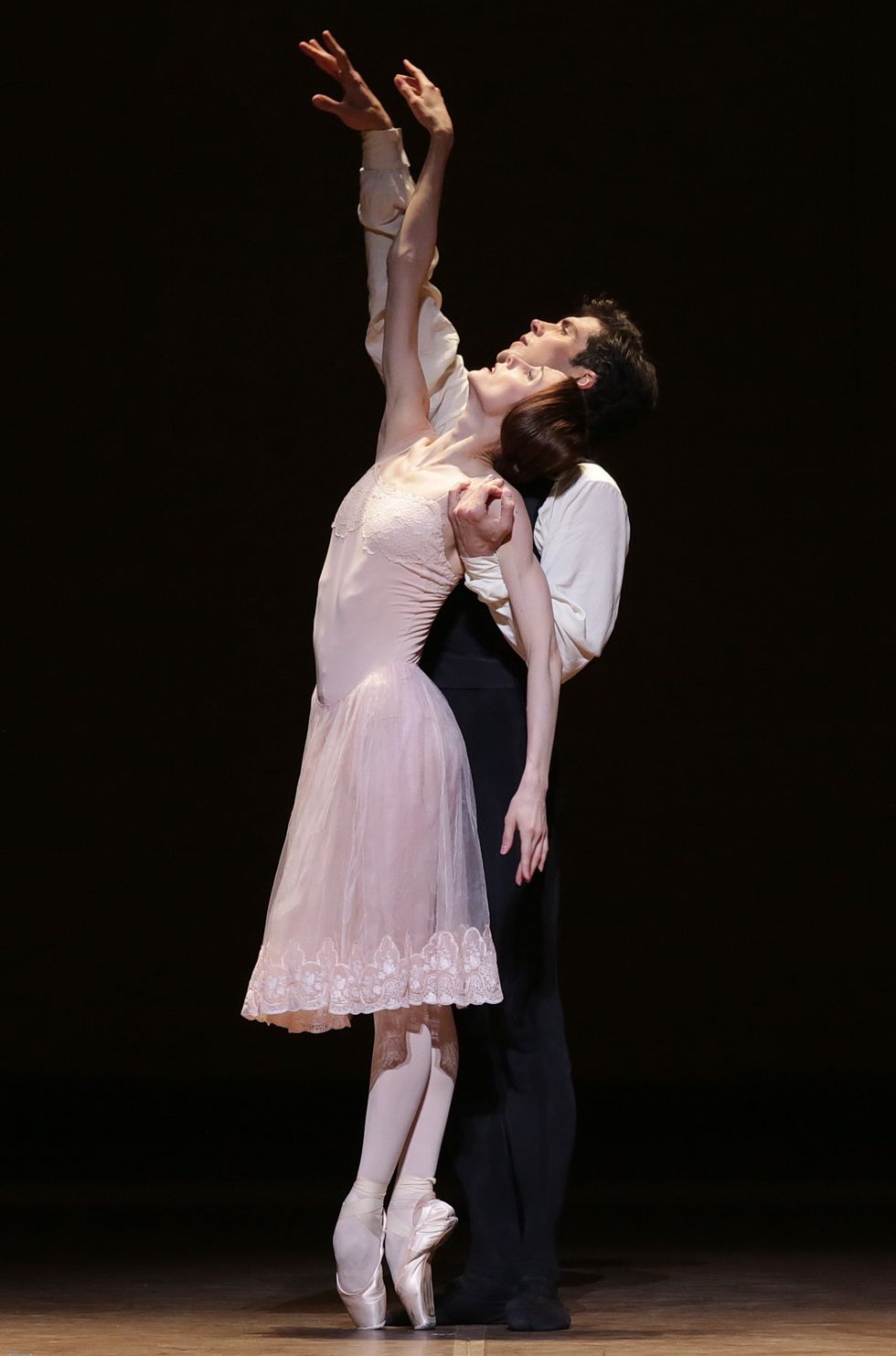 Zakharova balances in sous-sus en pointe, arching back as she reaches her right arm up, as Bolle stands behind her, supporting her under one arm while mirroring her reach with the other.