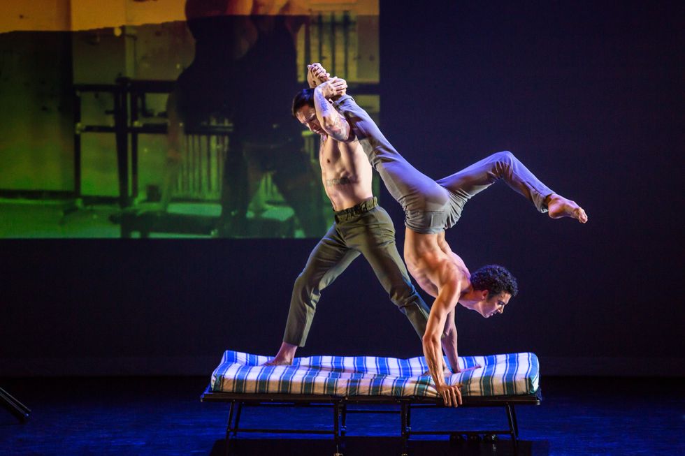 Two shirtless men balance on a narrow cot. One is in an inversion, one foot bending over his head. The other lunges facing away from him, holding the other man's foot behind his head.