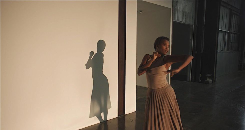 Shauna Davis, a young Black woman with short cropped hair, dressed in a beige tank top and pleated skirt, looks over her shoulder as she leans into one hip, arms following the motion. Her shadow shows clearly on the white wall beside her.
