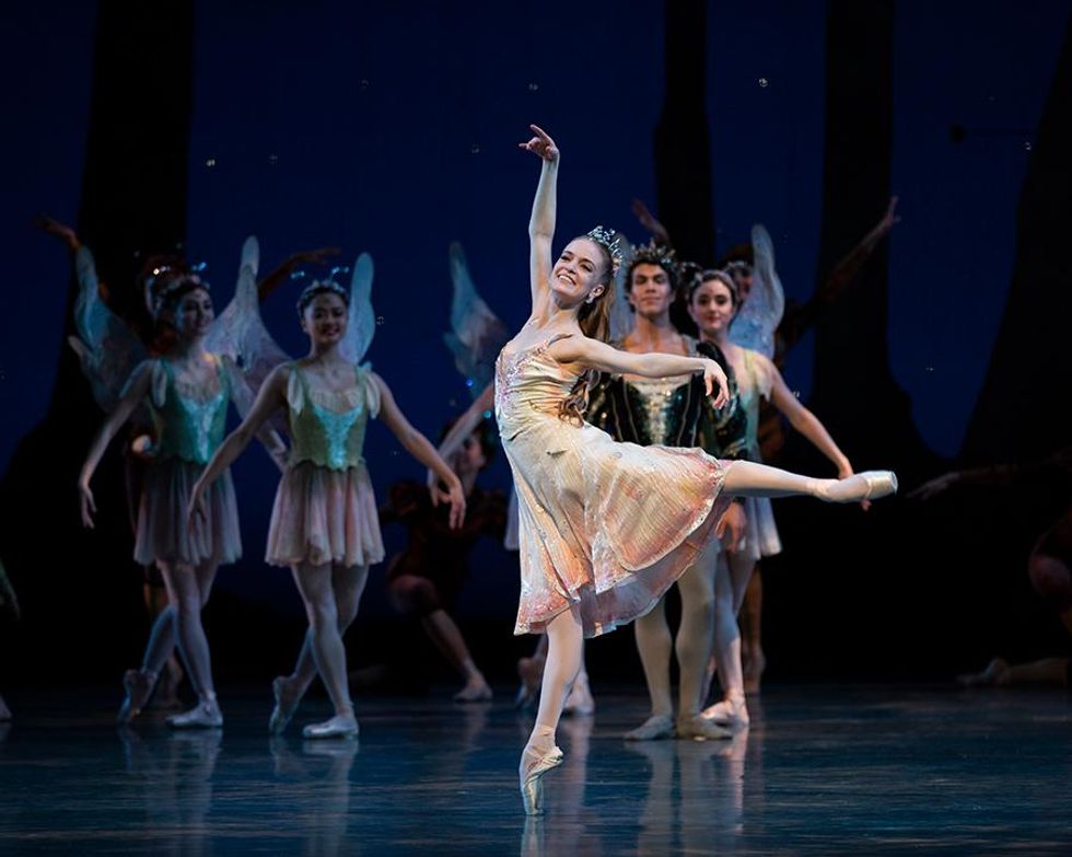 Sasha De Sola, a white woman costumed in a pale yellow dress, tiara, pink tights, and pointe shoes, smiles radiantly at the audience as she balances in first arabesque en pointe, working arm overhead in fifth.