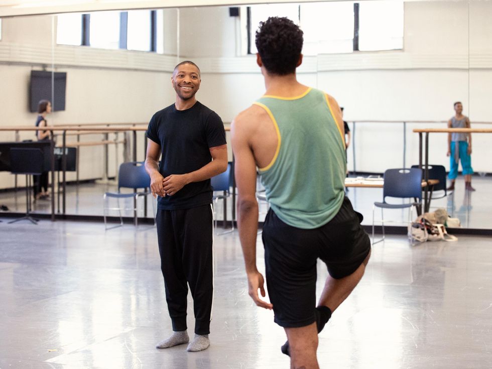 Kyle Abraham stands at the center of a large, sunlit ballet studio, smiling at a male dancer whose back is to the camera. Both wear sweats, t-shirts, and socks.