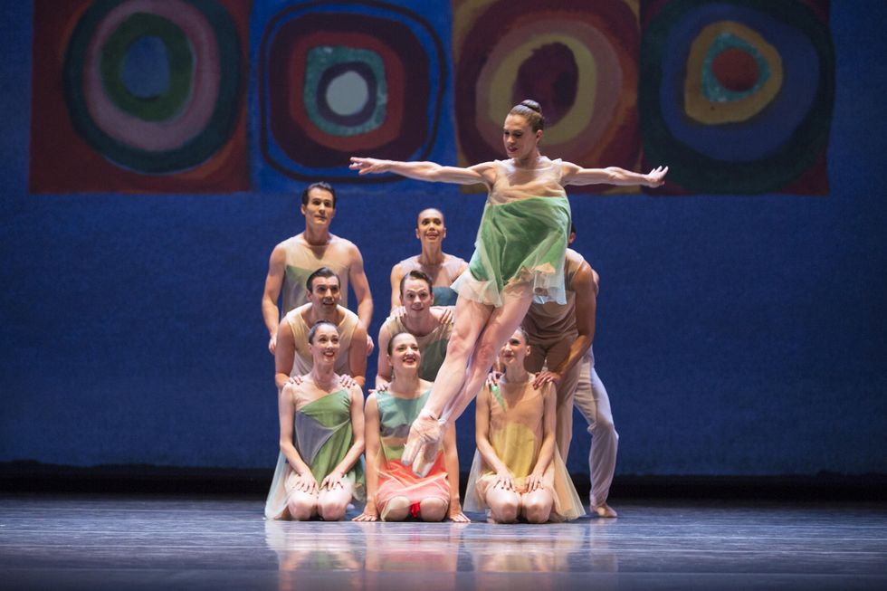 A female dancer in a flowy, colorful tunic assemblu00e9s in an open fifth, arms stretched to her sides. Upstage, a cluster of nine dancers arranged in a neat square, the front row kneeling, watch with bright smiles.