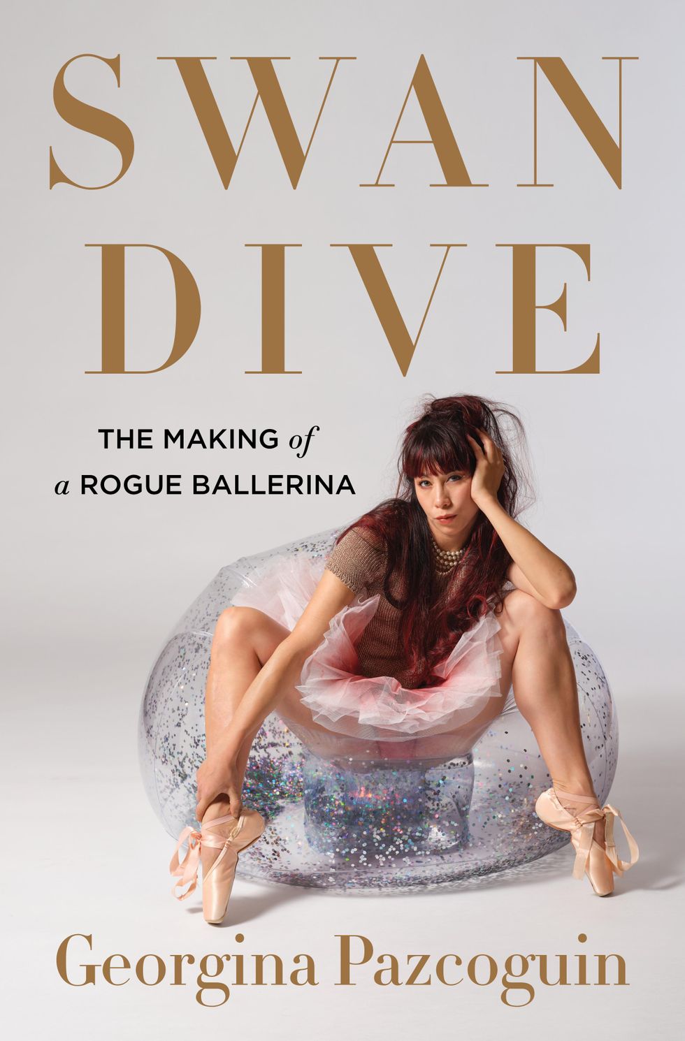 Against a grey backdrop, Georgina Pazcoguin sits in a glittery inflatable chair in bare legs, incorrectly tied pointe shoes, a girlish pink tutu and pearls. Her hair is down and she holds her head in one hand. In bronze text, the cover reads, "Swan Dive: The Making of a Rogue Ballerina. Georgina Pazcoguin."