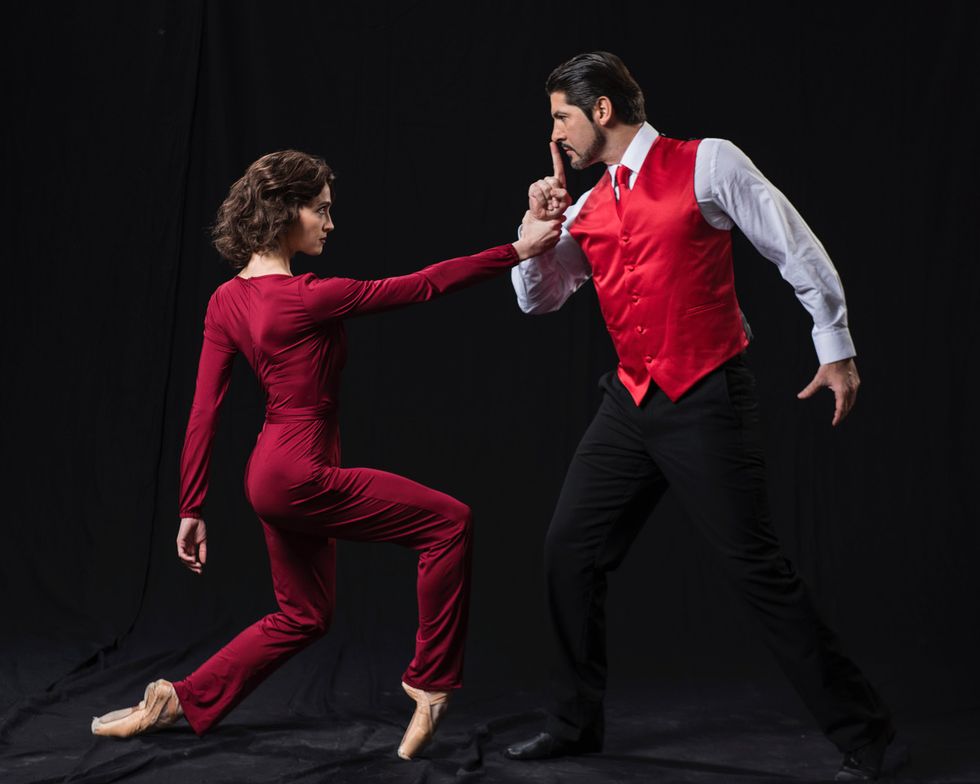 A woman in a red jumpsuit in pointe shoes balances in a parallel forced arch fourth position. Her eyes are intent on the man's whose wrist she is grabbing. Dressed in a matching red vest and tie, he leans toward her, holding a finger over his lips.