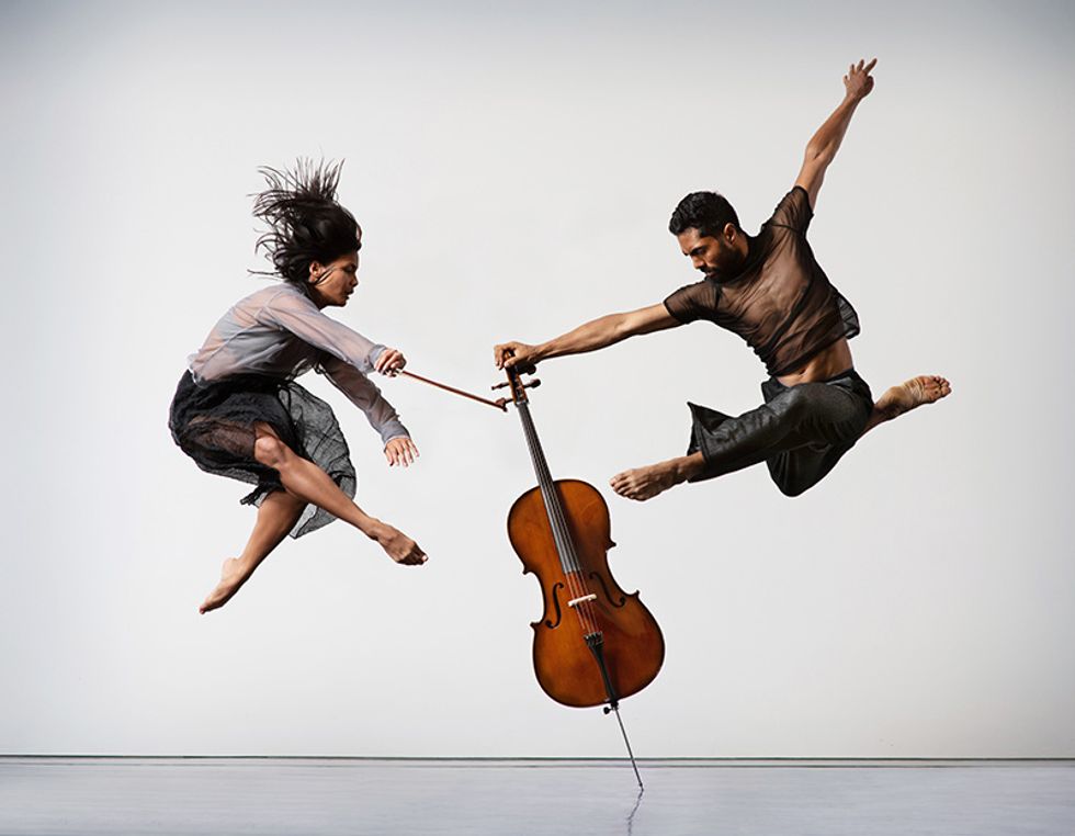 Two dancers are caught mid-jump, both legs folded underneath them. The male dancer balances a cello against the floor, while the female dancer brings a bow toward the strings.