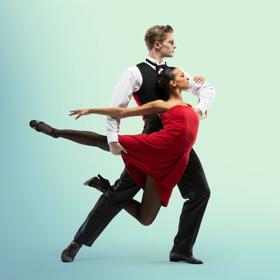 A male dancer wearing most of a suit holds a woman in a red dress in a fish pose, her bent knee tucked between his legs.
