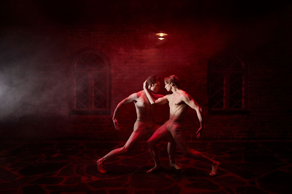 In a shadowed, smoky, red-lit space, two shirtless male dancers lunge toward each other, mirroring one another as they hold the back of the other's head and stare into each other's eyes.