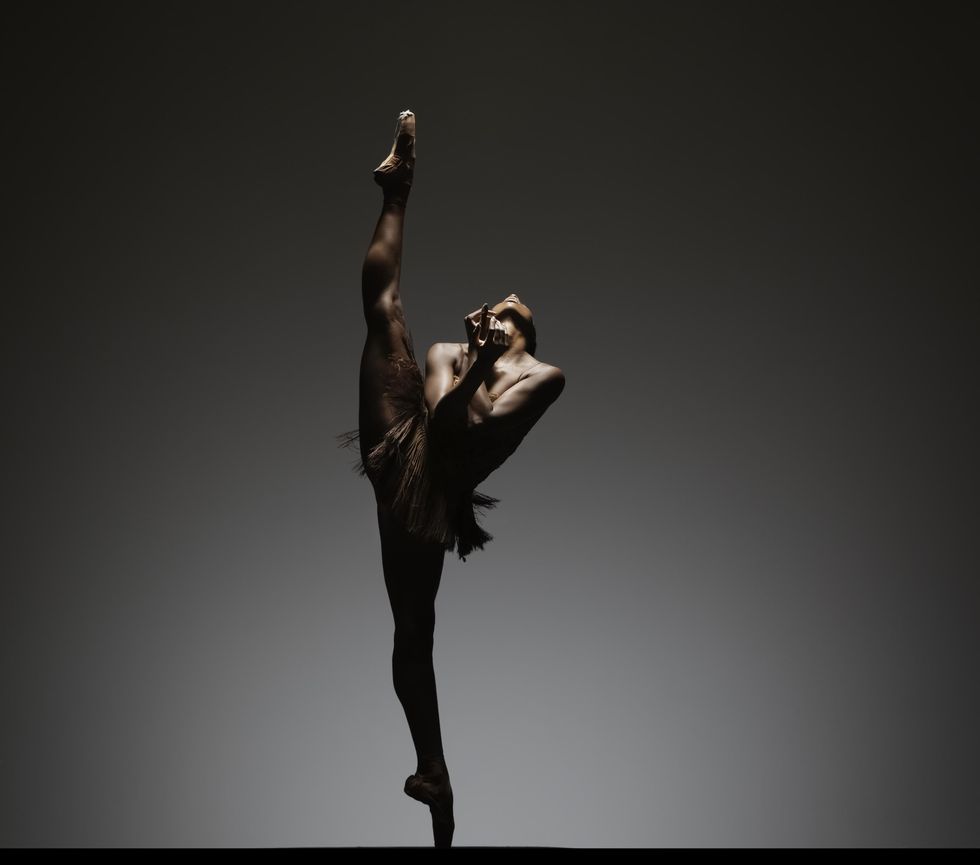 Against a grey background, a woman in pointe shoes and a short, tasseled dress balances on relevu00e9 in a side extension that stretches almost to 180 degrees. Her arms cross and fold at the wrists in front of her chest. Her head is tipped all the way back to gaze at the ceiling.