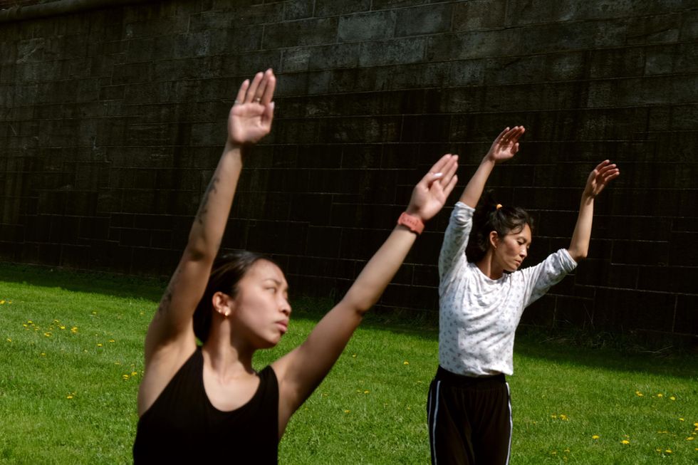 On a field of bright green grass, hemmed in by dark grey bricks or tiles, two dancers in greyscale rehearsal clothes stretch their arms overhead and lean lightly to their left, gazes downward.