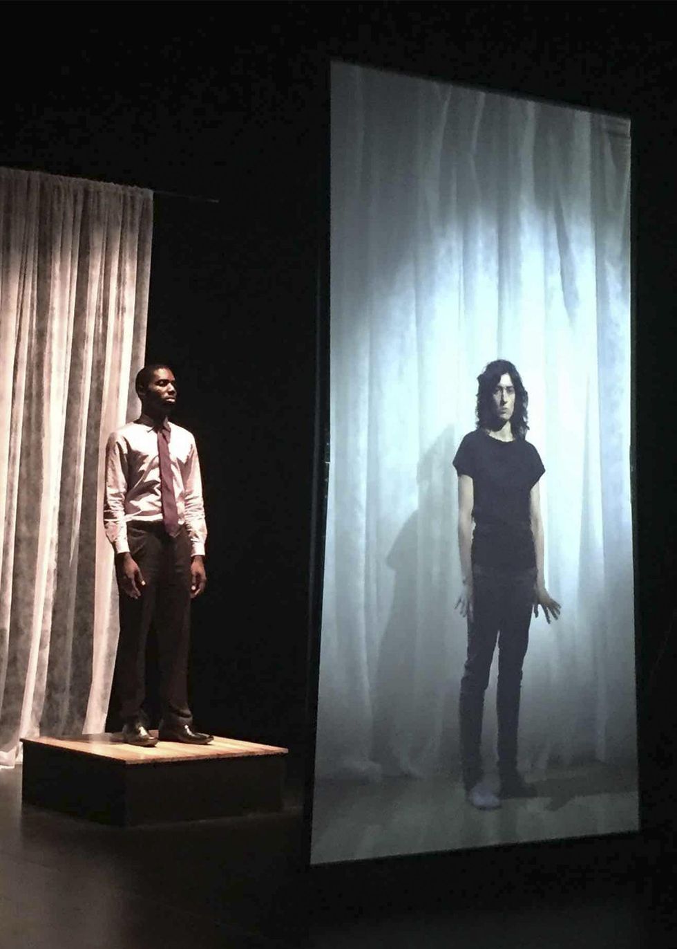 A man in a suit and tie, minus the jacket, stands on a foot-high platform, arms by his sides. He watches the life-size image of a dancer projected onto a screen in front of him. The dancer also stands in a neutral position, arms by their sides, but their fingers flare out to the sides as they look at the camera.