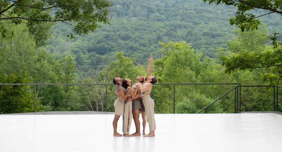 Five dancers wearing shades of beige and grey cluster together on a white stage with a see of green foliage behind. They hold each other's shoulders and look upwards as one dancer reaches a splayed hand overhead.