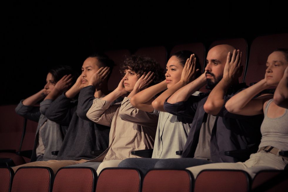 Six dancers dressed in shades of beige and gray sit in a row of red and black seats in a theater. All stare straight ahead, hands over their ears and elbows away from their torsos.