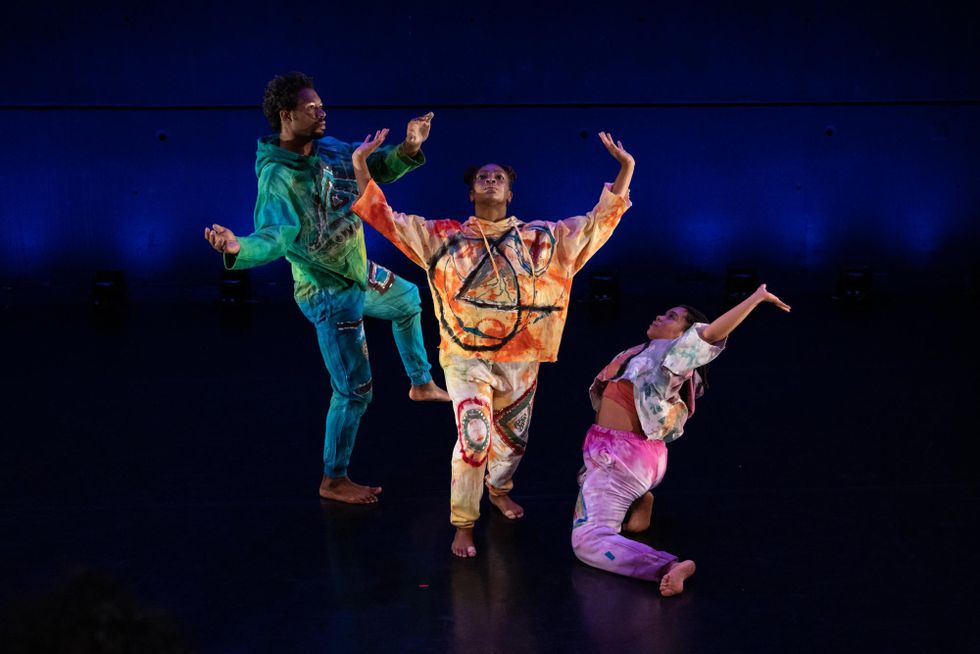 Three Black dancers wearing hoodies and sweatpants in bright colors, with patterns reminiscent of tie-dye, pose together on a blue and black stage. One balances meditatively on one leg; at the center, another raises her palms and eyes towards the rafters; a third kneels, looking up at the second as she lunges upstage.