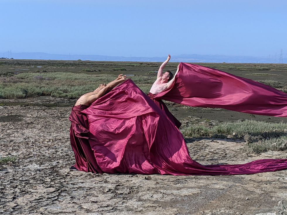 The torsos of two topless dancers emerge from billowing crimson fabric, the color rich and bright against the scrubland and hazy blue sky behind them.