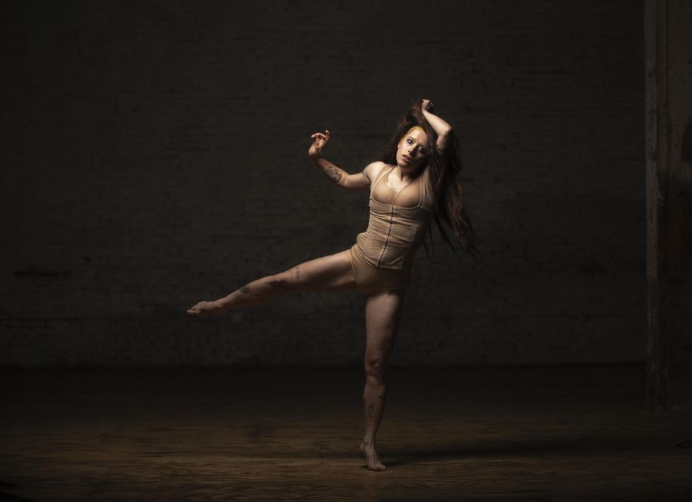 A dancer dressed in a flesh-tone leotard reminiscent of a corset balances precariously on relevu00e9, working leg in a parallel side attitude. They pull a long, flowing brown wig away from their scalp as they stare, wide-eyed and alarmed, at the camera.