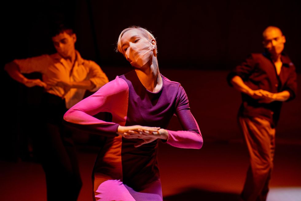 A woman clothed in bright pink and purple and a pale mask that matches her skin color tips her head to one side as she uses one palm to press her other hand down. Two other dancers mirror the motion in the blurred, shadowy background.