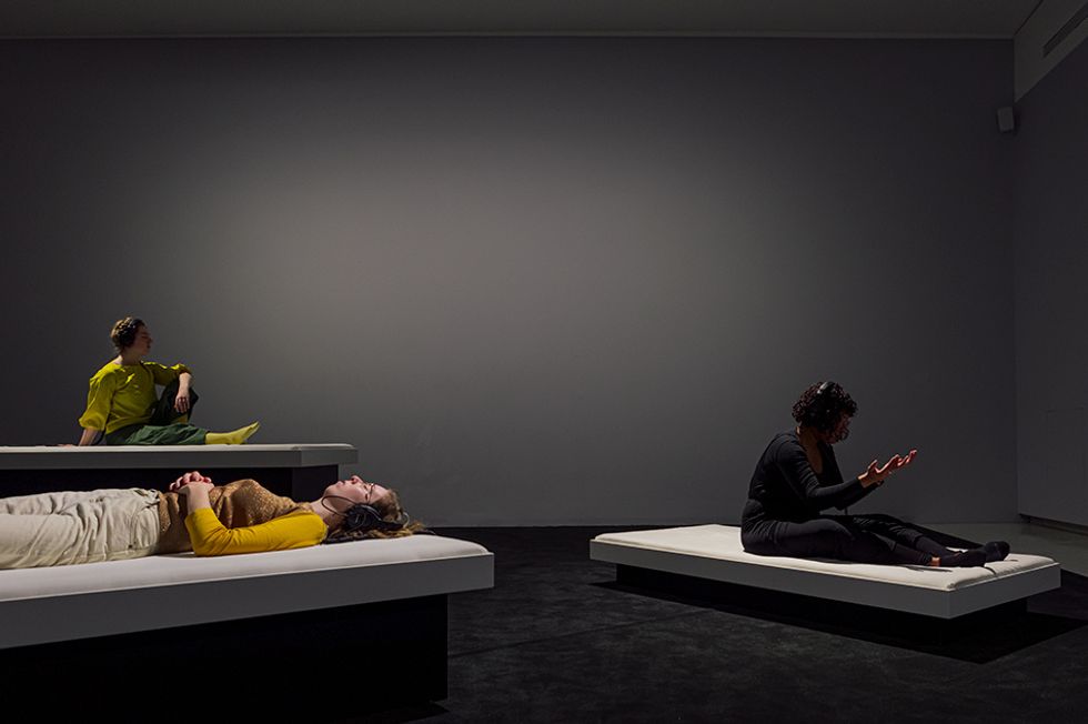 Three people sit or lie on white platforms raised to various levels above the ground, following physical cues from the headphones they wear.