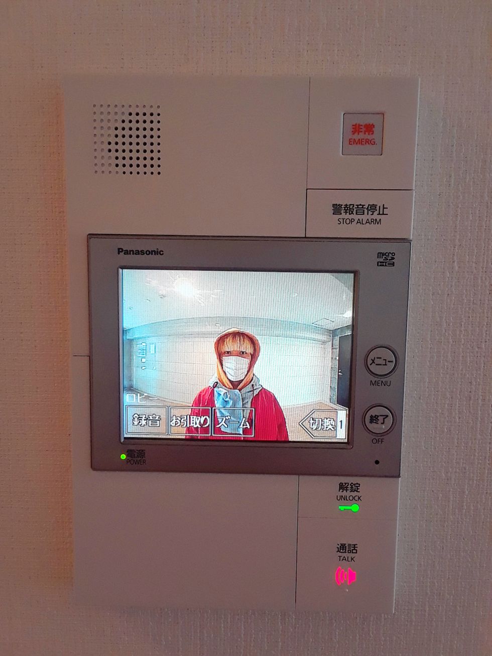 The video monitor on a door buzzer shows Mei Yamanaka looking into the camera from a hallway, wearing a voluminous red top, a medical mask, and a hood.