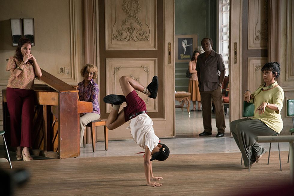 Edlison Manuel Olbera Nu00fau00f1ez, portraying a young Carlos Acosta in Yuli, does a handstand in sneakers and a baseball cap in the middle of an ornate studio. Two ballet teachers, an older pianist and his father look on with bemusement.