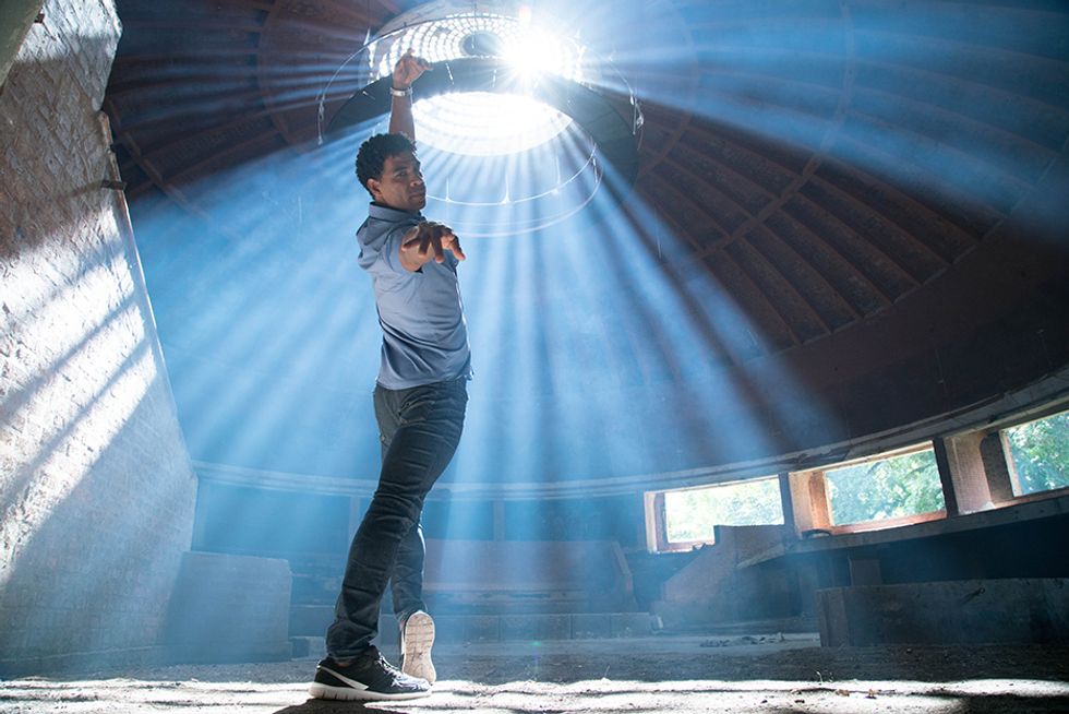 In a still from Yuli, Carlos Acosta, wearing jeans and a button down, looks over his shoulder to the camera from a forced-arch lunge. Sunlight drenches the abandoned building from a circular skylight.