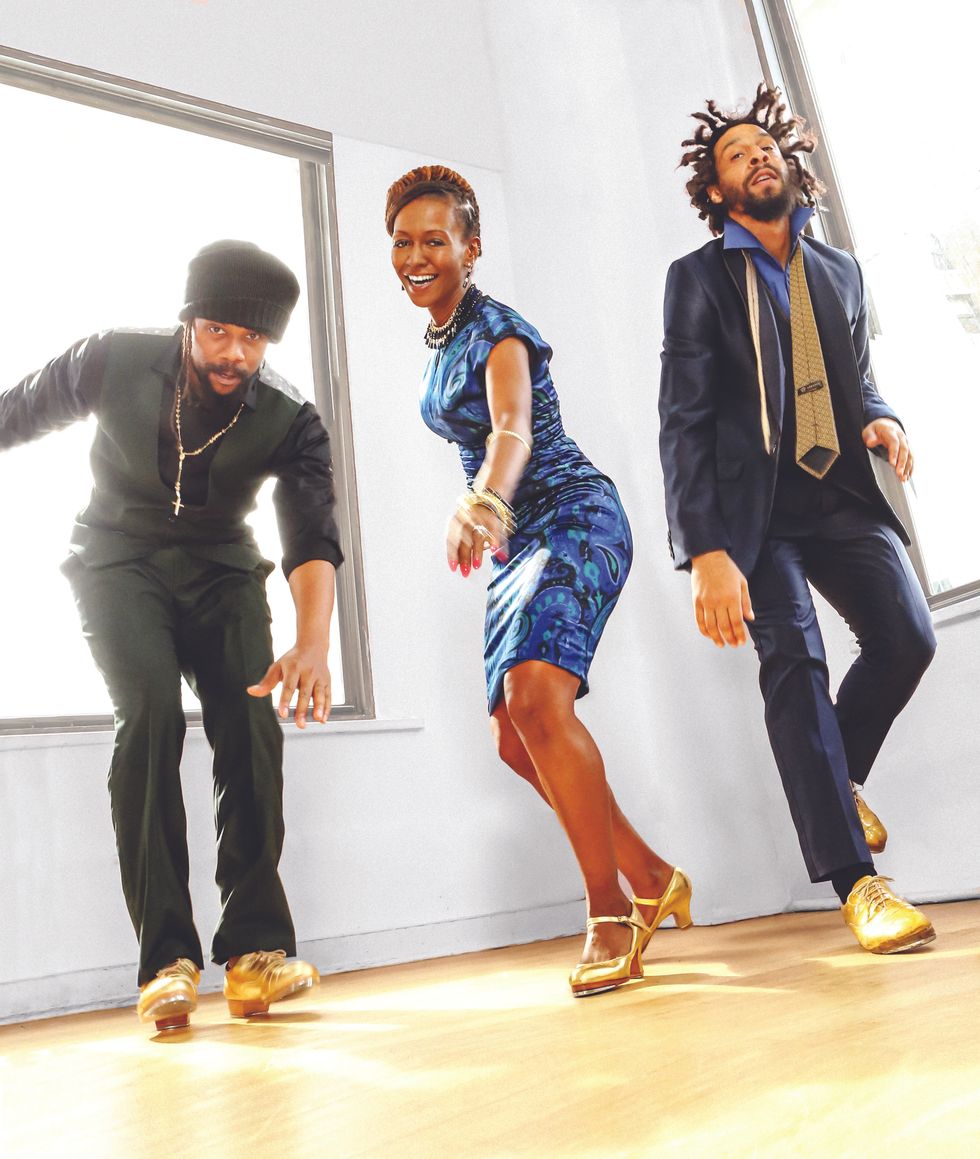 Derick K. Grant and Jason Samuels Smith, dressed in black and blue suits, and Dormeshia in a blue patterned dress, dance together in a sunlit corner, gold tap shoes shining.