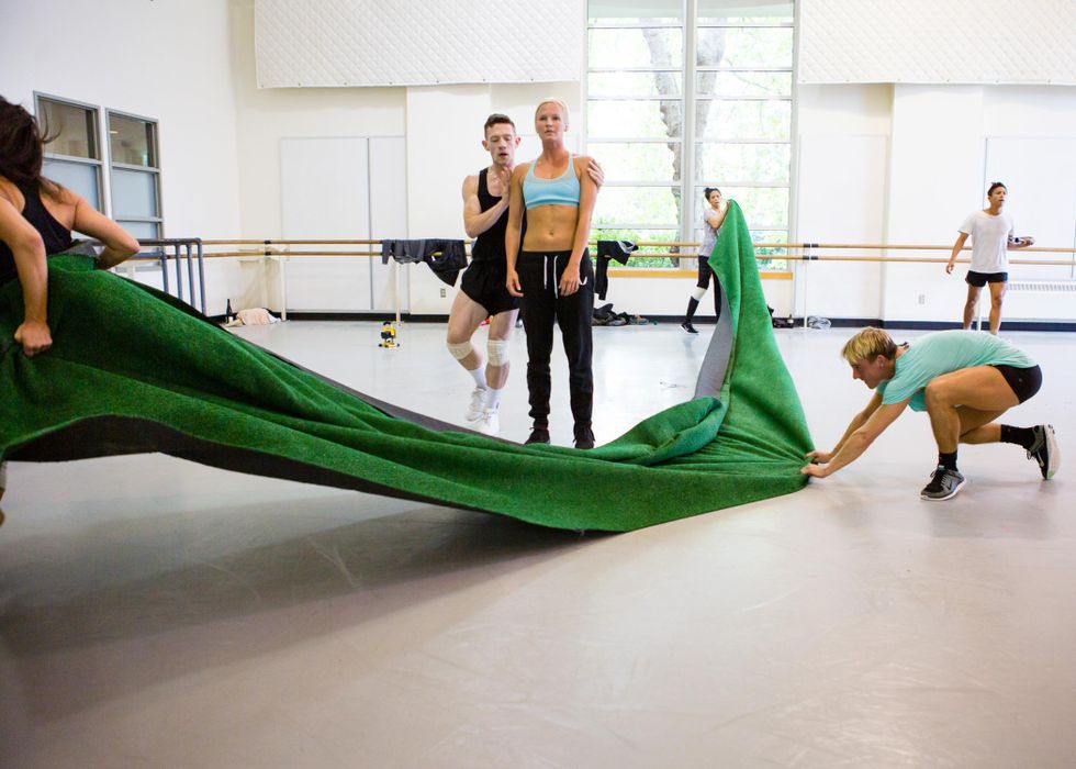 In an airy rehearsal studio, three dancers manipulate a long, thin length of green fabric that stretches from upstage to downstage. Two dancers stand just to one side of it, one grabbing the other by the shoulders as she stands neutrally, staring ahead.