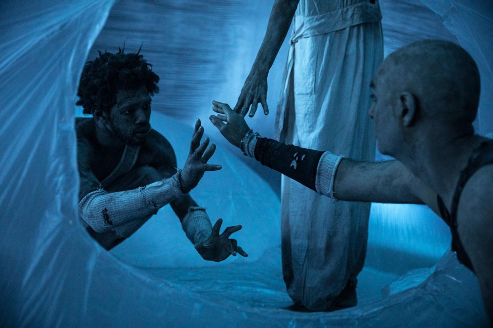 In a blue-lit space covered in plastic, two grimy figures in beat up clothing reach cautiously toward one another. A third figure reaches their hand down toward them, visible only from the waist down.