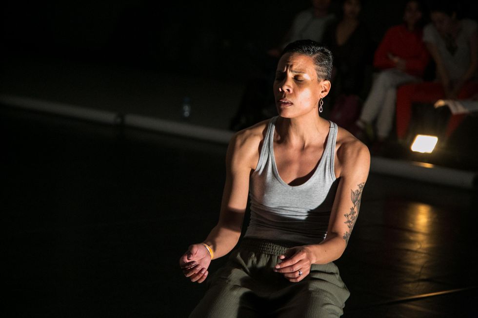 Cara Hagan closes her eyes as she kneels, hunching forward slightly as her arms, bent at the elbows and pulled toward her ribcage, begin to expose the underside of her wrists. Her brow is furrowed; her hair is pulled back; she wears a grey tank top and olive green trousers. A black tattoo of a flower is visible on one arm.