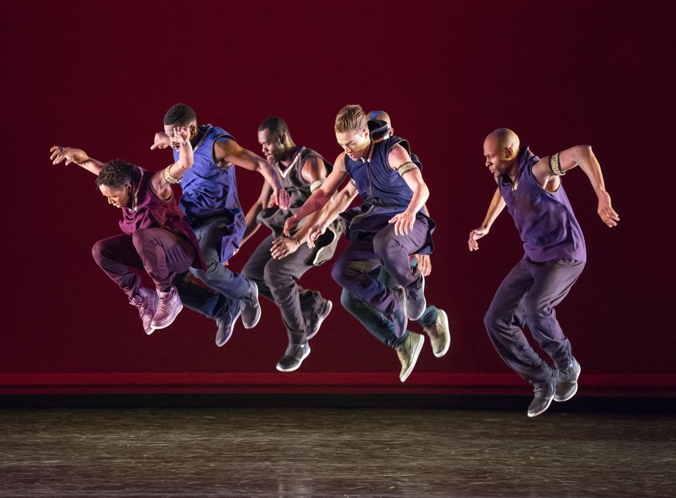 Six dancers dressed in shades of blue, purple and black are caught mid-air, knees pulling up towards chests, arms loosely raised to the sides, gazes cast downward.