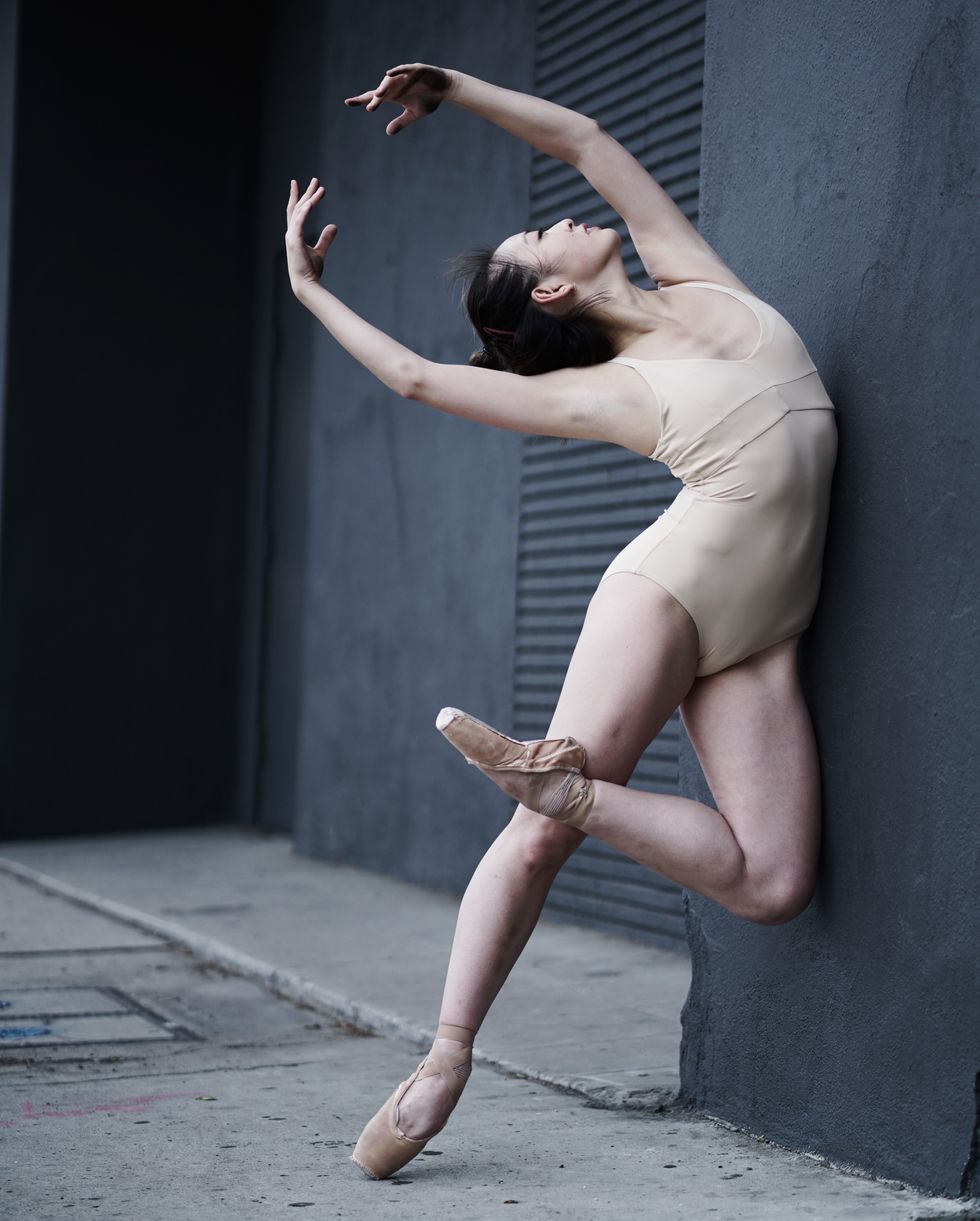 A dancer in nude leotard in a passe on pointe leans against a grey building