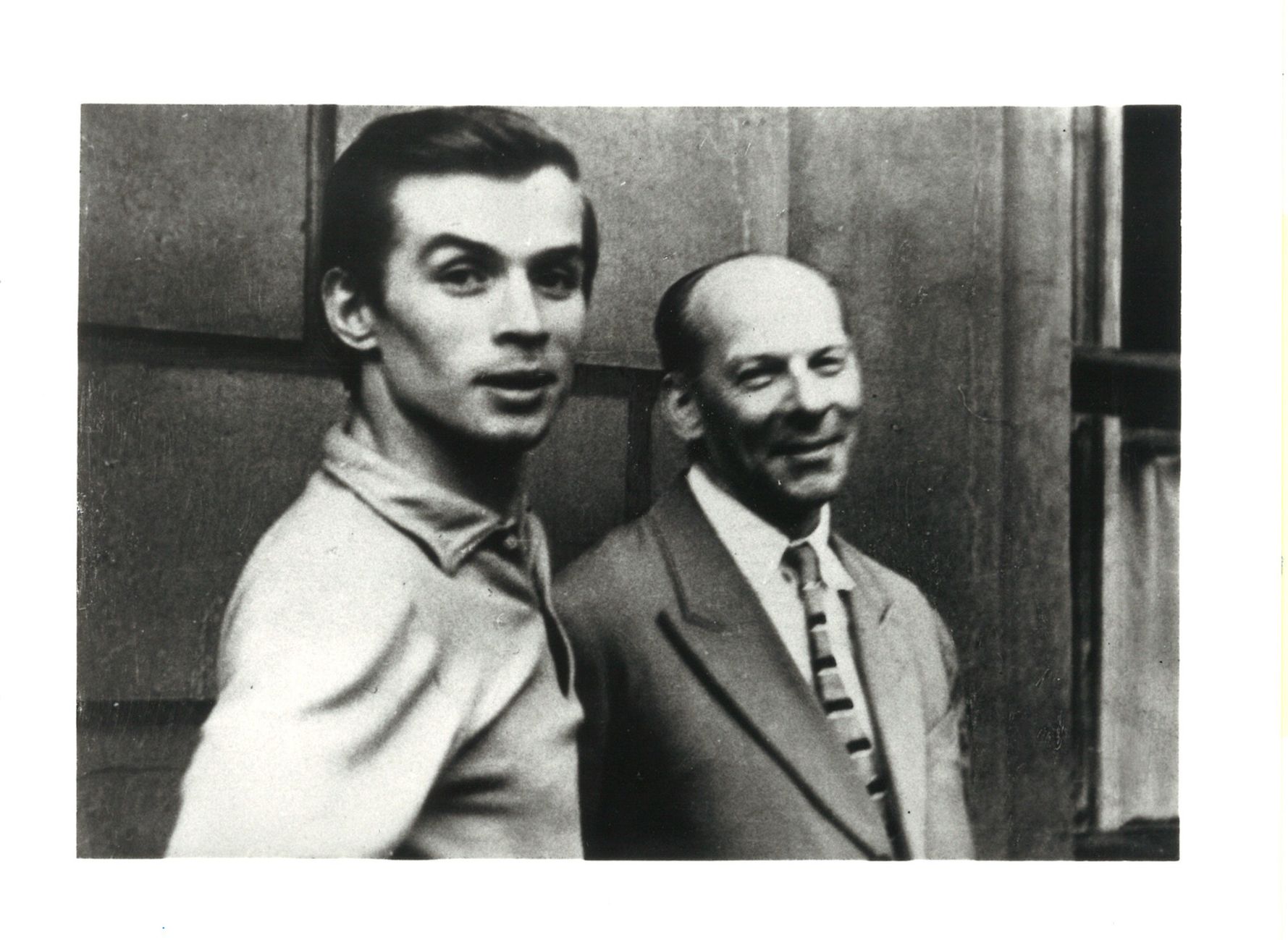 A black and white photograph of a young Rudolf Nureyev and his teacher, Alexander Pushkin, here smiling and wearing a suit and tie.