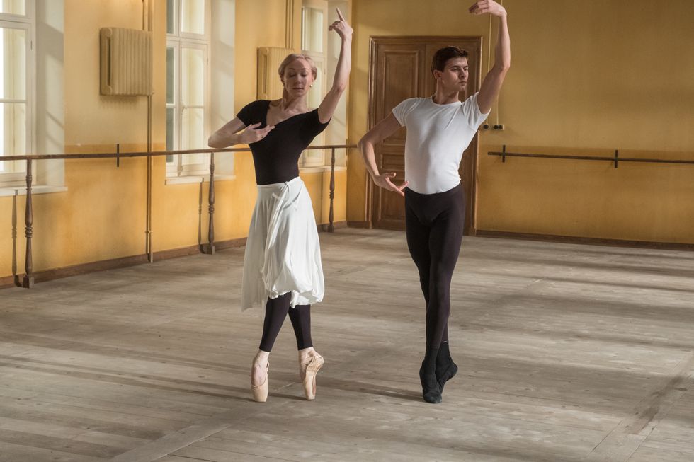 In the White Crow, Natalia Dudinskaya, an older, blonde ballerina, and Rudolf Nureyev rehearse in the studio. Both wear practice clothes and are posed in releve, arms in fourth position.