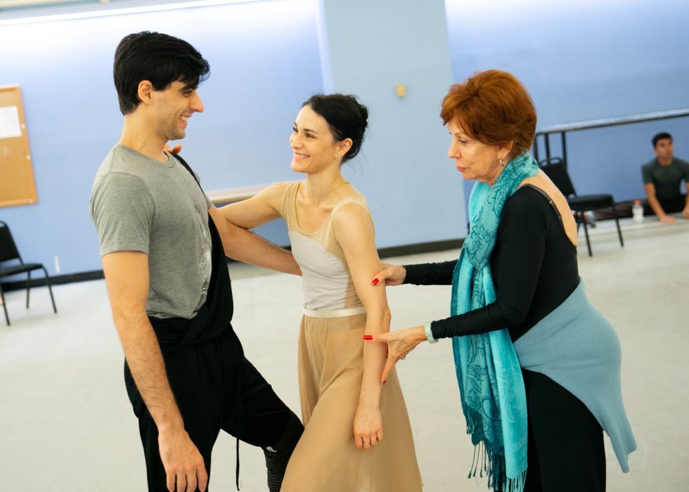 Calero-Alonso, with a colorful blue scarf draped over her shoulders, uses both hands to adjust Lane's arm, which is loose at her side. Lane laughs with Ribagorda; they face each other, standing loosely with their left legs in forced arch, upstage arms linked around each other's shoulder and waist.